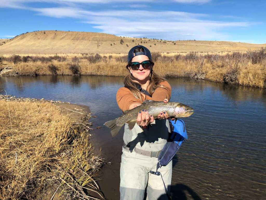 Lindsay is standing in a stream holding a fish.